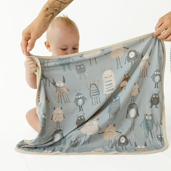 A baby is partially hidden behind a large light blue cloth towel or blanket printed with cartoon monsters, held up by two adult hands against a clean white background.