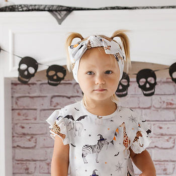 A toddler stands barefoot in front of a white fireplace mantel decorated for Halloween, wearing a white short-sleeve dress with a brown and black animal print and a matching headband. Ghost decorations, black webbing, skull garlands, and pumpkins are visible in the room.