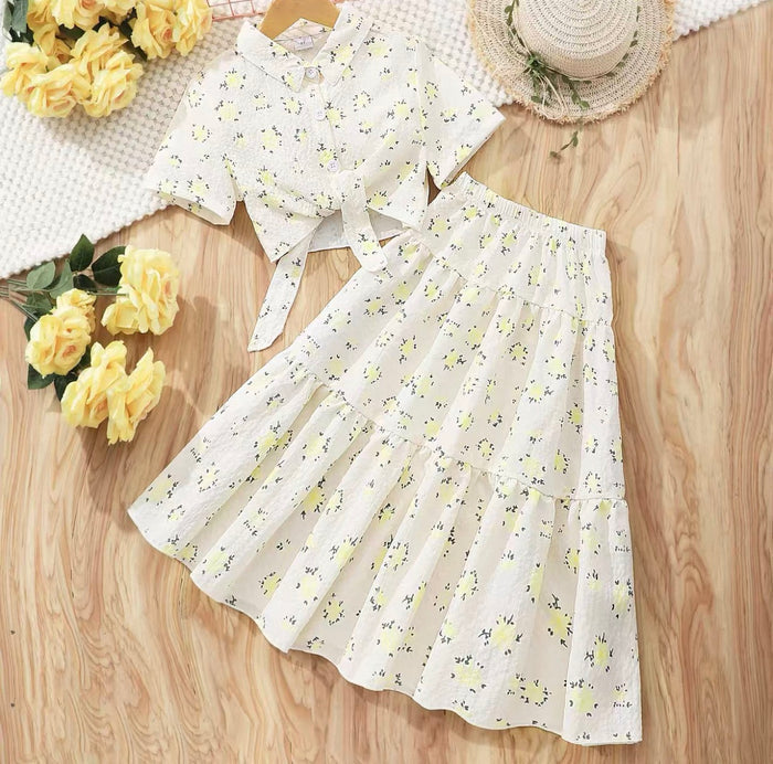 White floral outfit with a skirt and top on a wooden surface with yellow flowers and a hat.