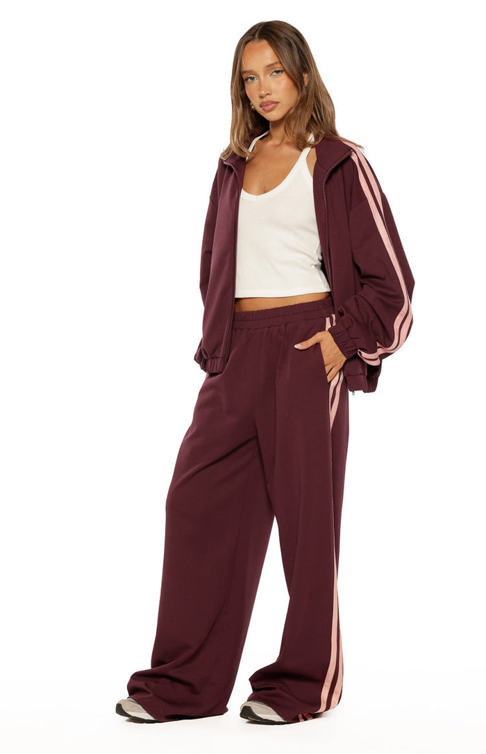 Burgundy wide-leg track pants with pink side stripes, showing the relaxed fit and elasticated waistband