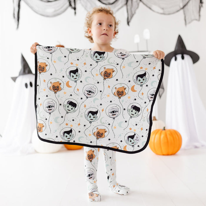 A young child stands holding up a white blanket or sheet with a repeating Halloween vampire balloon pattern, in a decorated Halloween-themed room with pumpkins and ghost props.