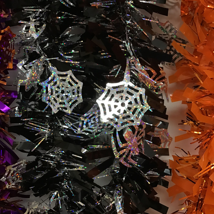 Holographic spider and web Halloween decorations hanging on a black tinsel tree, with orange tinsel visible on the right side.