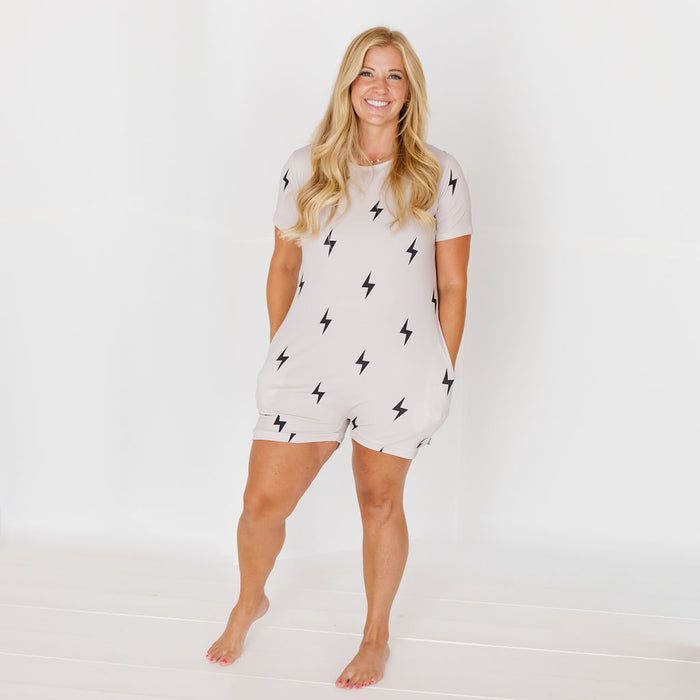 A woman modeling a light beige short-sleeve romper with a black lightning bolt pattern, standing against a white studio background.