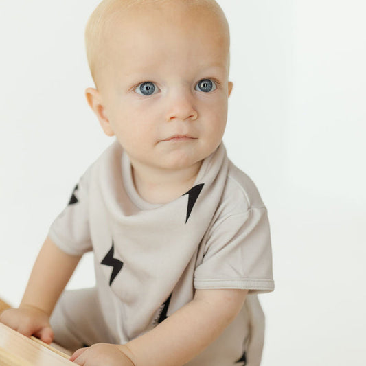 A baby with light hair and blue eyes wearing a beige short-sleeve romper with black lightning bolt prints, sitting on a wooden surface against a white background.