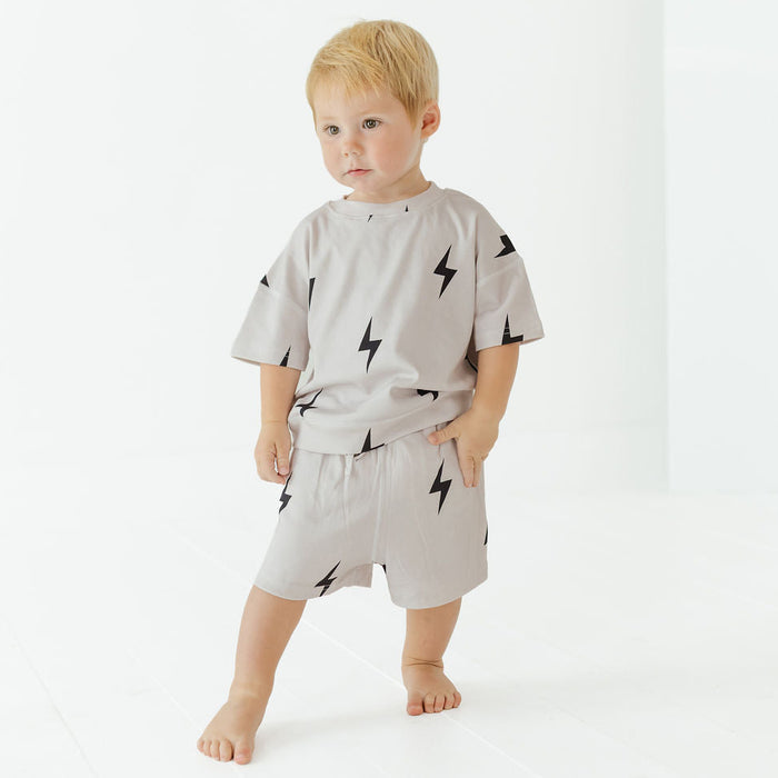 A toddler standing barefoot in a bright white studio, wearing a light gray short-sleeve t-shirt and matching shorts with a black lightning-bolt print.