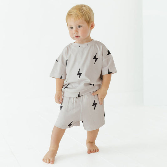 A toddler standing barefoot in a bright white studio, wearing a light gray short-sleeve t-shirt and matching shorts with a black lightning-bolt print.