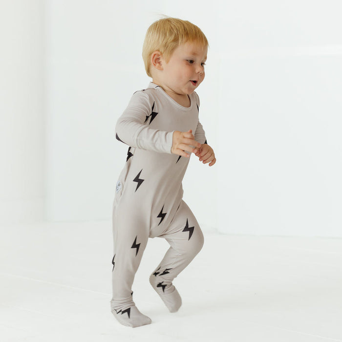 A toddler with blonde hair wearing a light gray long-sleeve footed sleeper with black lightning bolt prints, captured mid-step against a clean white studio background.