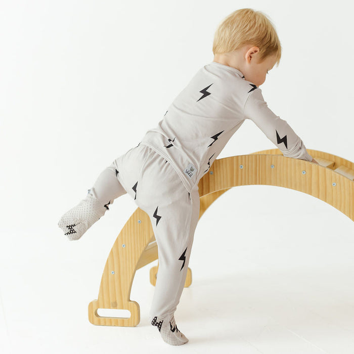 A toddler wearing a light gray long-sleeve pajama set with black lightning bolt prints is climbing a curved wooden play structure on a white studio background.