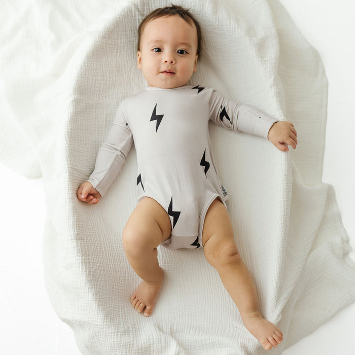 A baby lying on a white muslin blanket, wearing a light gray long-sleeve bodysuit with black lightning bolt prints.