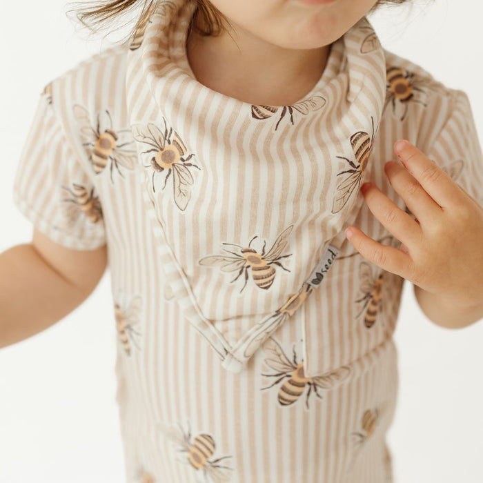 A close-up of a child wearing a short-sleeve bib or romper in beige and white vertical stripes with a printed bee pattern, photographed against a plain light background.