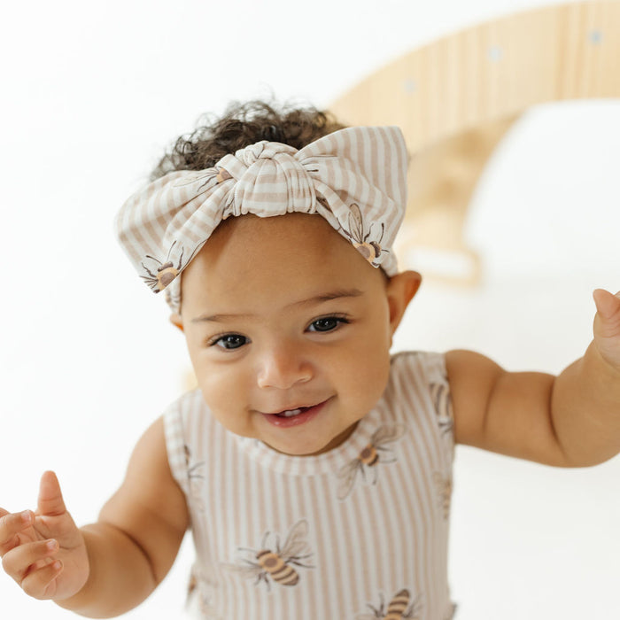 A baby wearing a beige and white striped bow headband and a matching sleeveless top with bee prints, smiling and reaching forward against a bright white background.