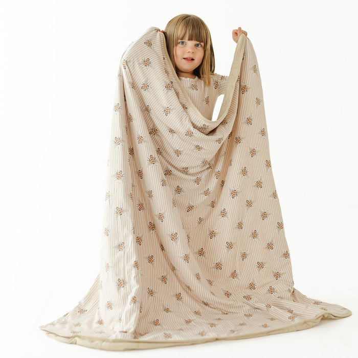 A young child with short blonde hair is wrapped in a large cream-colored blanket with a brown floral pattern, holding the top corners up against a plain white studio background.