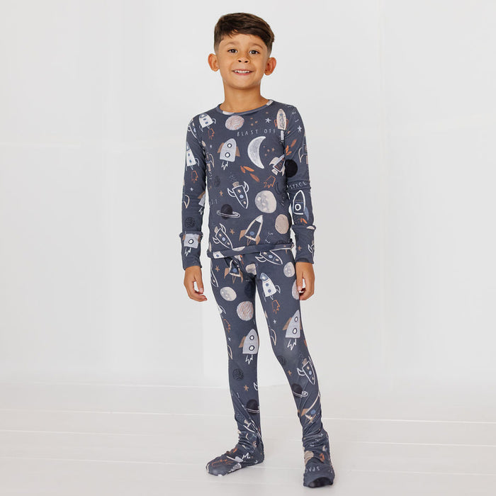 A young boy wearing a dark gray long-sleeve pajama set with a white and orange space-themed print of rockets, planets, and stars, standing against a plain white studio background.