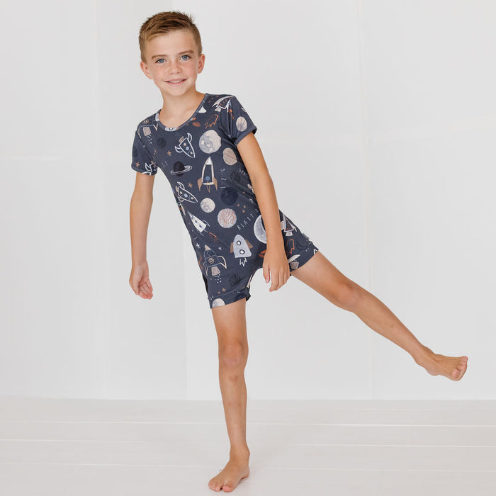A young boy wearing a short-sleeve pajama romper with a space-themed print of planets, rockets, and astronauts, posed against a clean white studio background.
