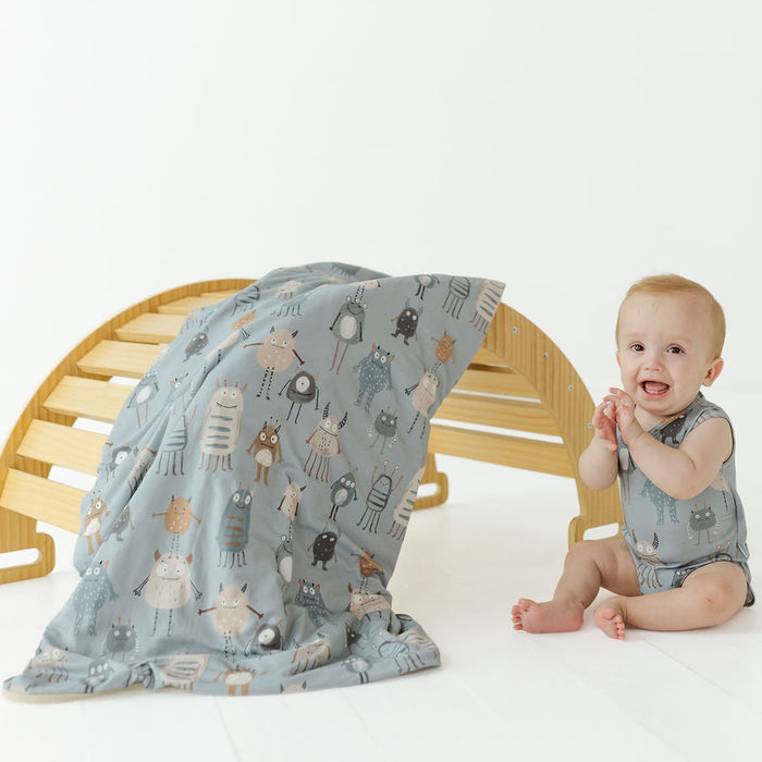 A baby wearing a matching light blue-gray romper sits beside a wooden bench draped with a matching patterned blanket. The fabric features a whimsical print of small cartoon animals in muted brown, beige, and gray tones.