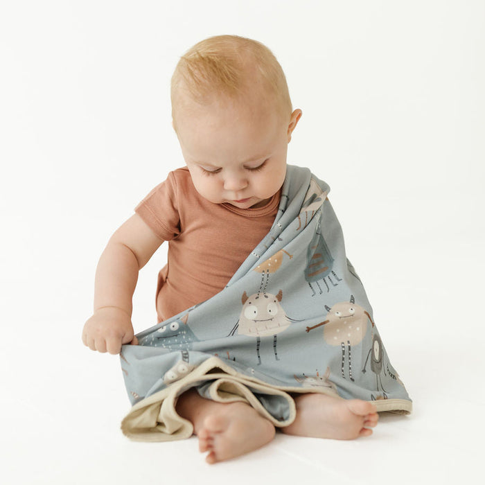 A baby sitting on a white studio floor wearing a dusty blue animal-print muslin swaddle wrap, paired with a terracotta short-sleeve bodysuit.