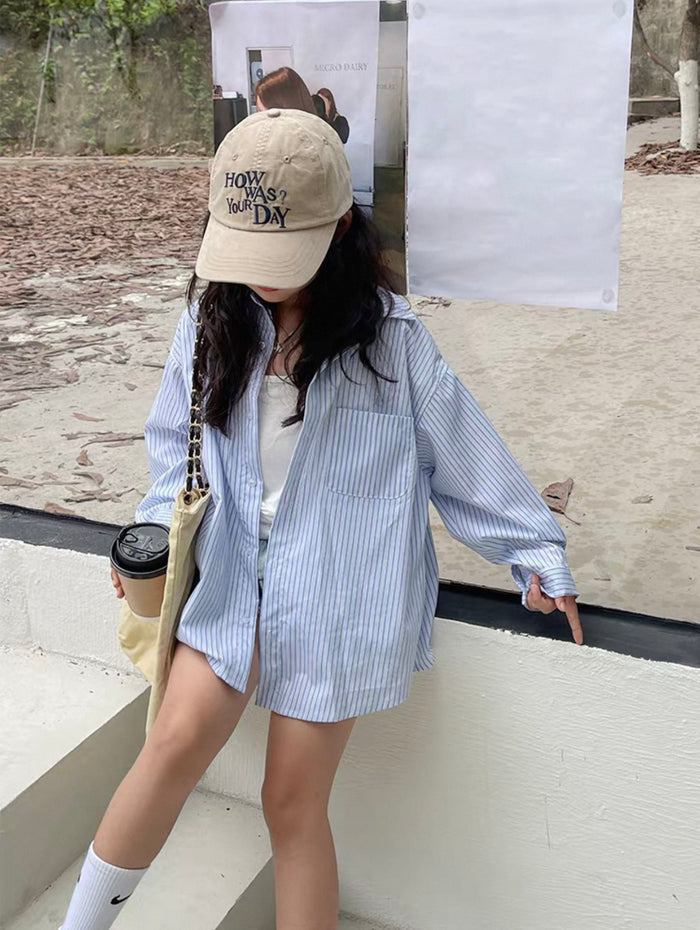 Person wearing a blue striped shirt and beige cap, holding a coffee cup outdoors.