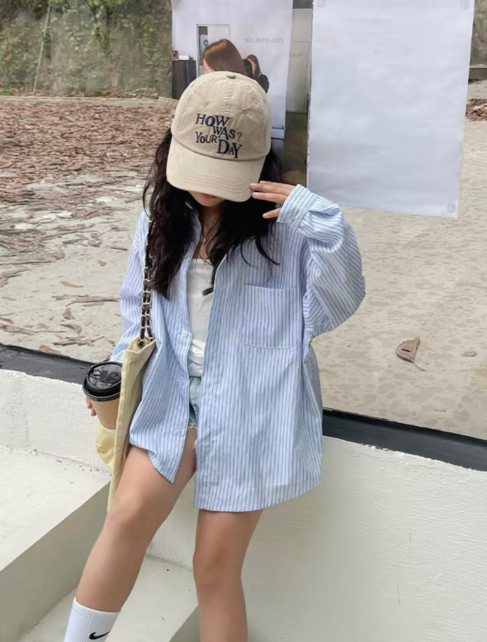 Person wearing a beige cap with text, blue striped shirt, and shorts, holding a coffee cup.
