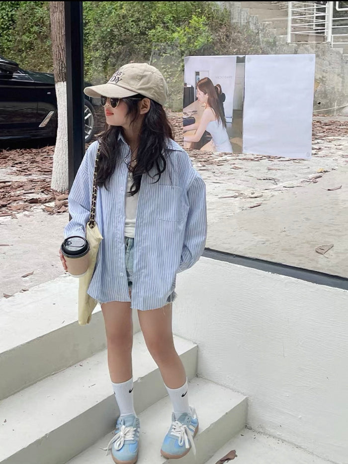 Woman standing on steps wearing a light blue striped shirt, denim shorts, and white sneakers.