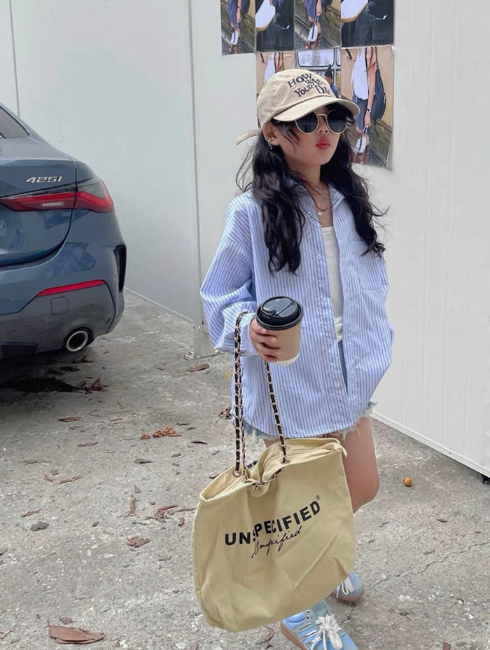 Person holding a beige tote bag with 'UNPEIFIED' branding, wearing sunglasses and a cap, standing outdoors.