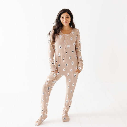 A woman modeling a beige footed pajama one-piece with an all-over white skull pattern, standing against a clean white studio background.