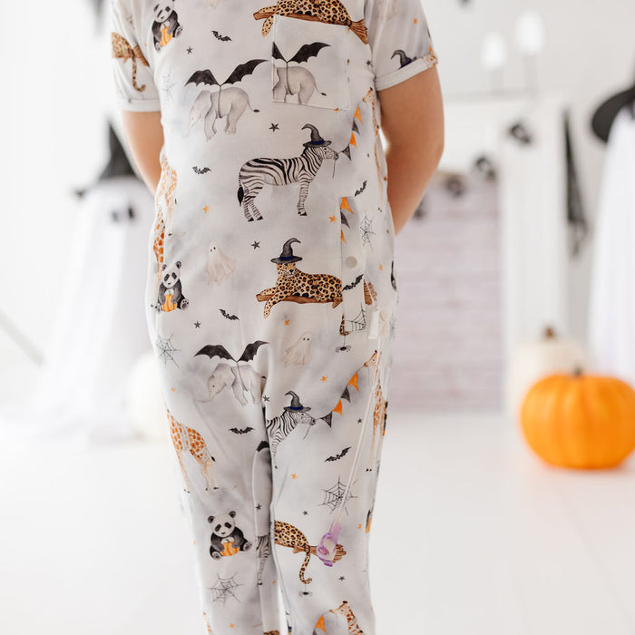 A child wearing a short-sleeve one-piece pajama or jumpsuit with a Halloween animal print, shown from the back in a bright indoor room with pumpkins and a ghost visible in the background.