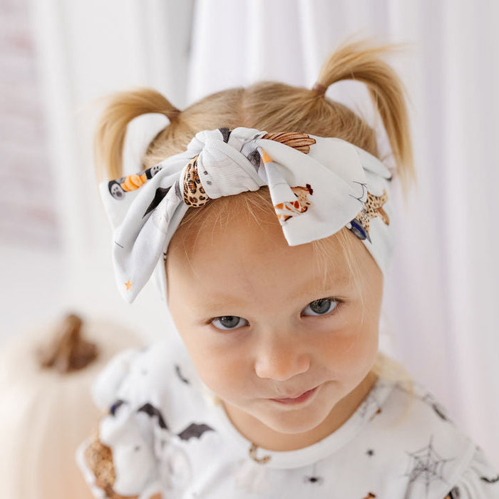 A toddler with blonde pigtails wearing a white fabric headband with a Halloween print and a matching white short-sleeve top, posed in front of white curtains.