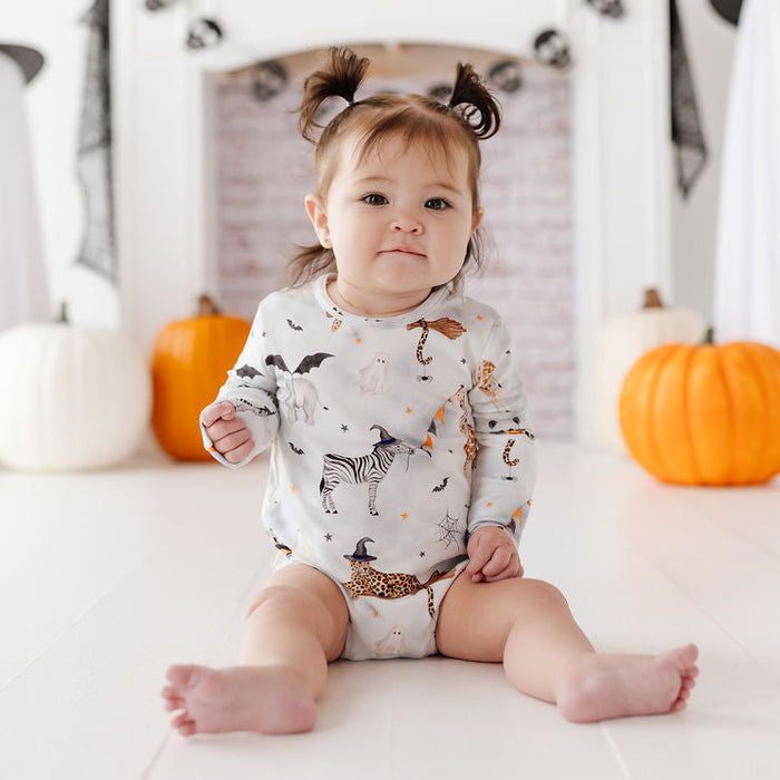A baby girl wearing a white long-sleeve onesie with a black and orange Halloween print sits on a white floor in front of a fireplace decorated for Halloween, with pumpkins and ghost props.