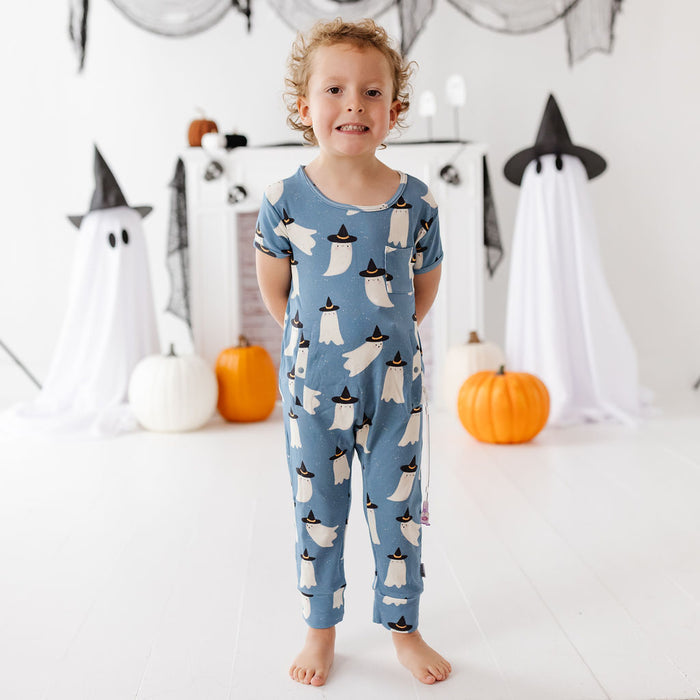 A young child stands barefoot in a white room decorated for Halloween, wearing a blue short-sleeve one-piece pajama with a repeating ghost pattern. Ghosts are white with black pointed hats, and pumpkins are placed on the floor around the child.
