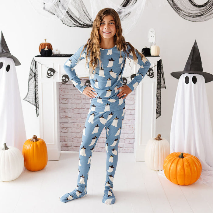 A young girl stands in a Halloween-themed room wearing a light blue long-sleeve pajama set with a white penguin print. She is posed in front of a white fireplace mantel decorated with black spiderwebs, black witch hats, white ghost decorations, and orange pumpkins.