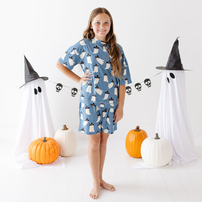 A young girl stands barefoot in a studio wearing a blue short-sleeve nightshirt or pajama top with a white ghost print. She is surrounded by Halloween decorations including two white ghost figures with black pointed hats, a skull banner, and orange and white pumpkins on a white floor against a white background.