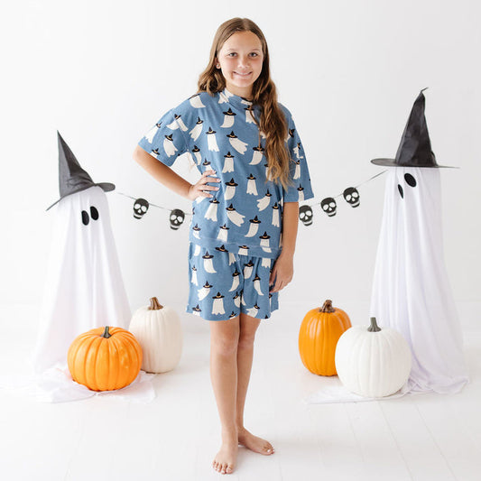 A young girl stands barefoot in a studio wearing a blue short-sleeve nightshirt or pajama top with a white ghost print. She is surrounded by Halloween decorations including two white ghost figures with black pointed hats, a skull banner, and orange and white pumpkins on a white floor against a white background.