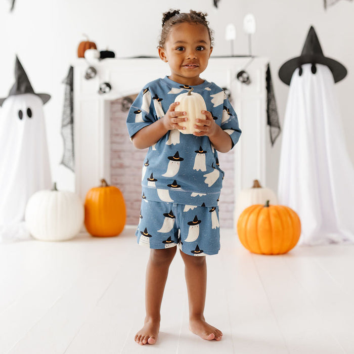 A young child stands barefoot in a Halloween-themed studio setting, wearing a blue pajama set with a penguin print and holding a small pumpkin. The background features white ghost decorations, black witch hats, pumpkins, and a white fireplace mantel.