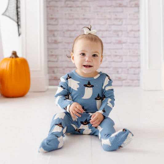 A baby sitting in front of a white fireplace mantel wearing a blue footed pajama set with a white ghost pattern. The scene is decorated for Halloween with pumpkins, ghost cutouts, and skull garland.