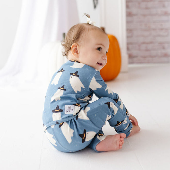A baby sitting on a white floor wearing a light blue one-piece pajama with a white ghost pattern, looking back over their shoulder in a styled room with a pumpkin and brick wall backdrop.