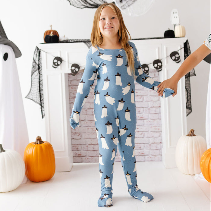 A young girl stands in a decorated Halloween room wearing a light blue one-piece pajama with a repeating white ghost pattern wearing black hats. An adult's arm is holding her hand on the right side.