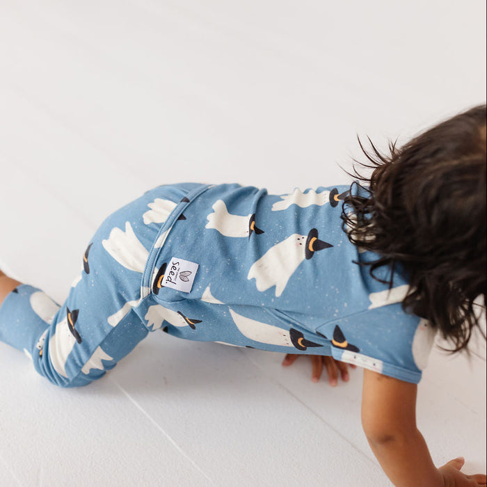 A baby crawling on a white floor wearing a light blue one-piece pajama with a white ghost pattern and black accents. The brand name 'Peachy' is visible on a small fabric tag on the back of the pajama.