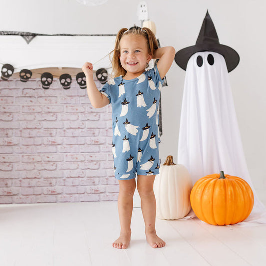 A young girl stands barefoot in a Halloween-themed room, wearing a light blue short-sleeve romper covered in a white ghost pattern. She is surrounded by fall decorations including a large white ghost, a paper witch hat, pumpkins, and a brick-patterned fireplace mantel.