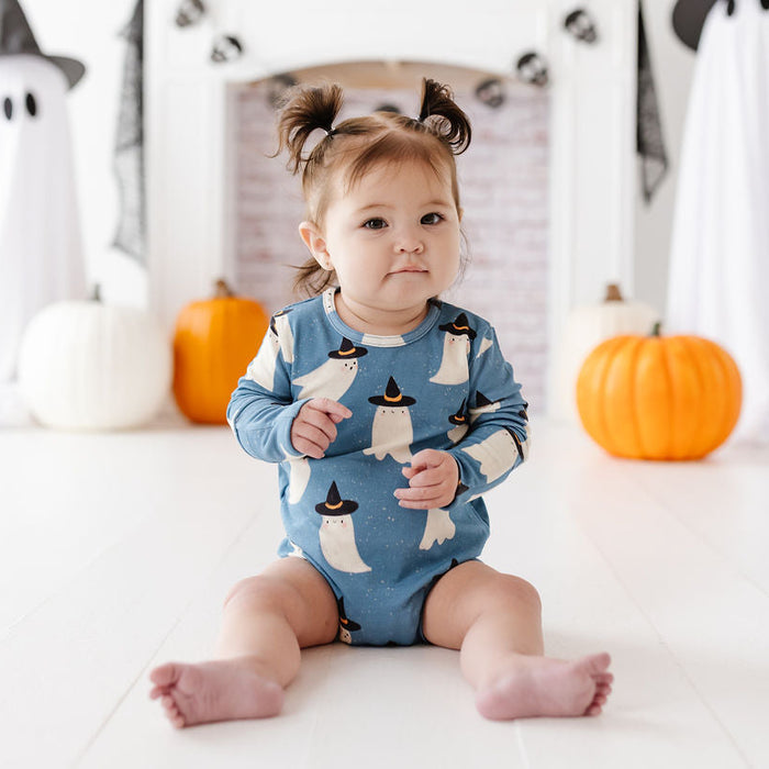 A baby sitting on a white floor wearing a blue long-sleeve romper with a penguin pattern, surrounded by Halloween decorations including pumpkins, ghost cutouts, and a fireplace backdrop.