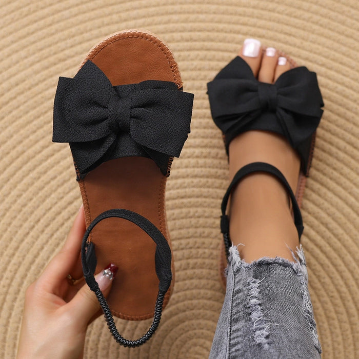 Black sandals with large bows worn on a textured beige surface