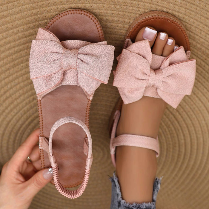 Pink sandals with large bows worn by a person on a textured brown surface