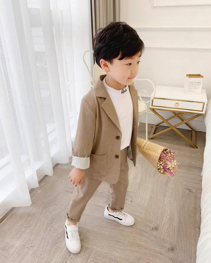 Child in a beige suit standing in a room with white curtains and a small table.
