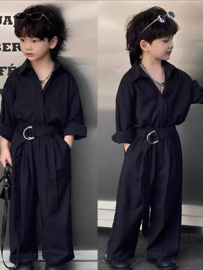 Child wearing a dark shirt and pants suit with a belt in front of a gray background