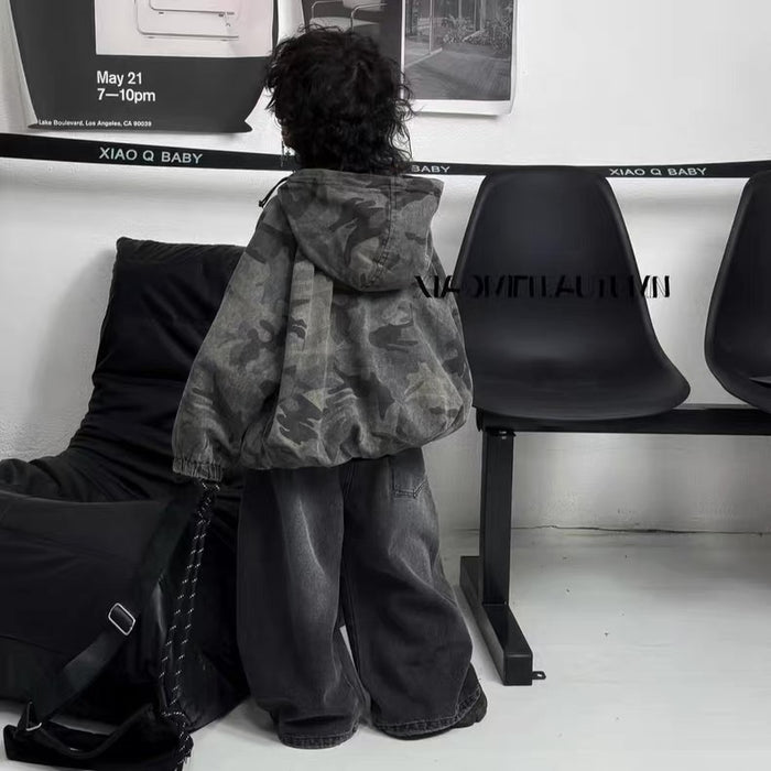 Person wearing a camouflage jacket and dark pants sitting on a black chair in a minimalistic room.