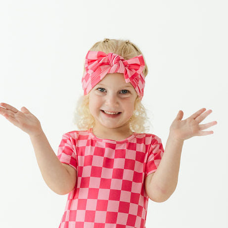 A young girl with blonde hair wearing a pink and white checkered short-sleeve swimsuit and a matching pink checkered headband, standing against a solid white studio background with her hands outstretched.