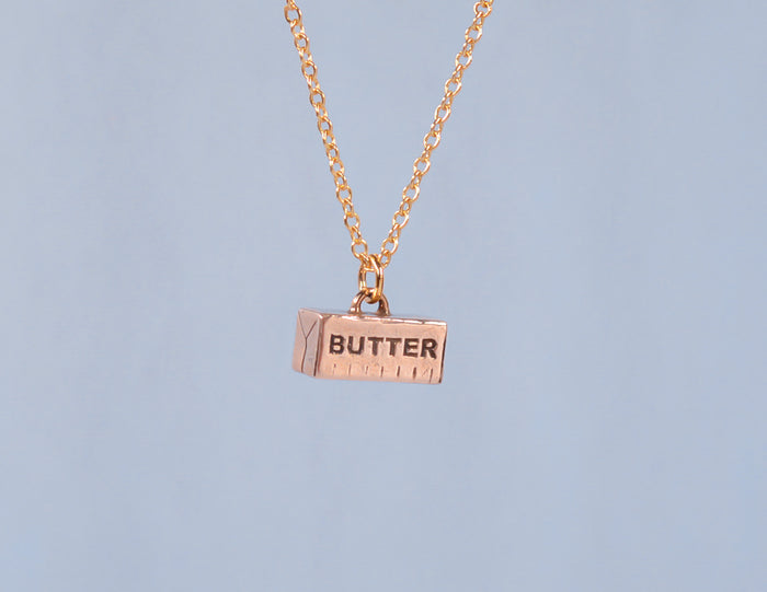 A bronze necklace in the shape of a stick of butter, with the word 'BUTTER' engraved on it, suspended on a 14k gold fill chain against a blue background.