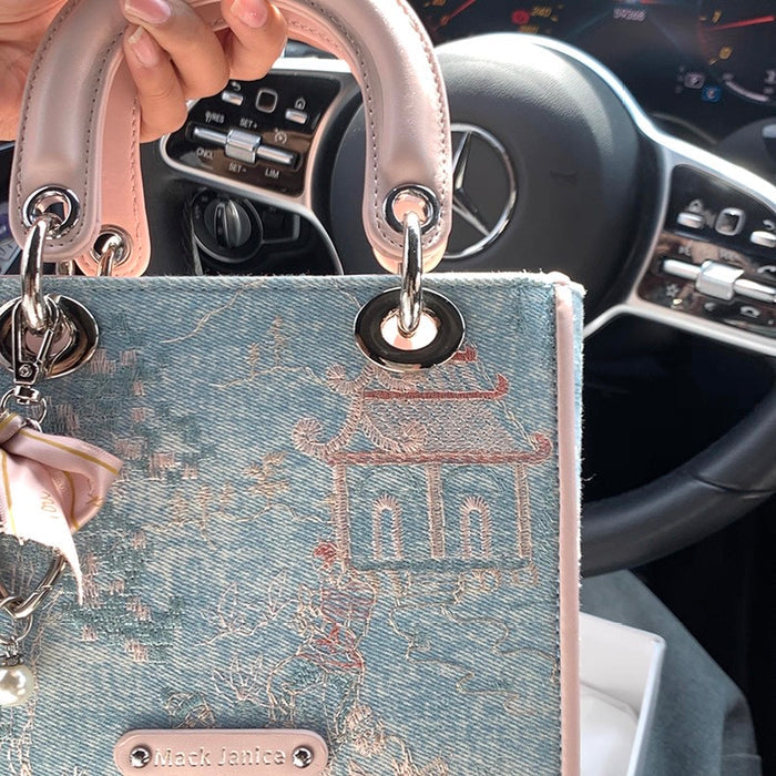 Handbag with intricate design held in front of a car's steering wheel and dashboard.