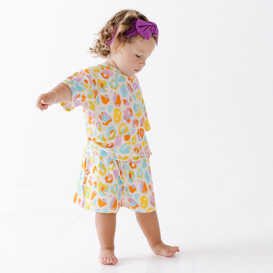 A toddler girl wearing a matching two-piece pajama set with a colorful abstract print, standing on a white studio floor. The set features a short-sleeve top and shorts in pastel shades of orange, yellow, pink, and blue on a white base.