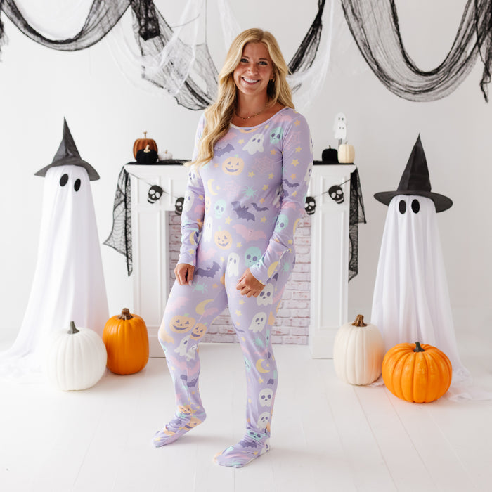 A woman stands in a Halloween-themed room wearing a light lavender long-sleeve pajama set with a cute ghost and moon pattern. The room is decorated with white ghost figures draped in black sheets, orange pumpkins, and black netting.