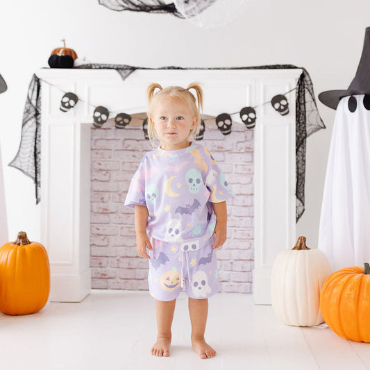A toddler stands barefoot in a Halloween-themed room, wearing a light lavender pajama set with a colorful print of ghosts, skulls, and moons. The background features a white fireplace mantel decorated with skull garlands, black spiderwebs, ghost cutouts, and orange and white pumpkins.